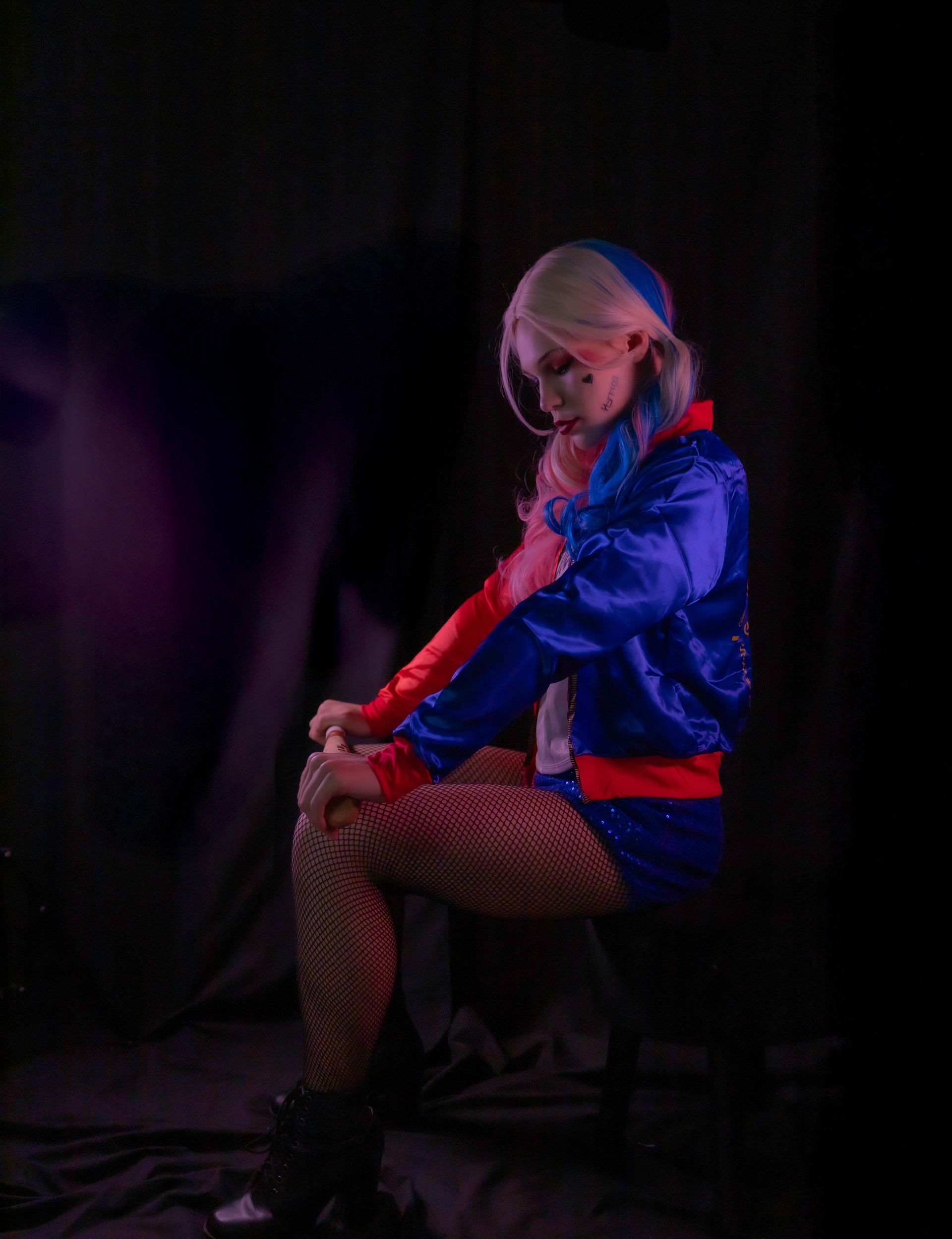 Harley Quinn cosplayer sits with head down, wearing blue and red jacket, fishnets.