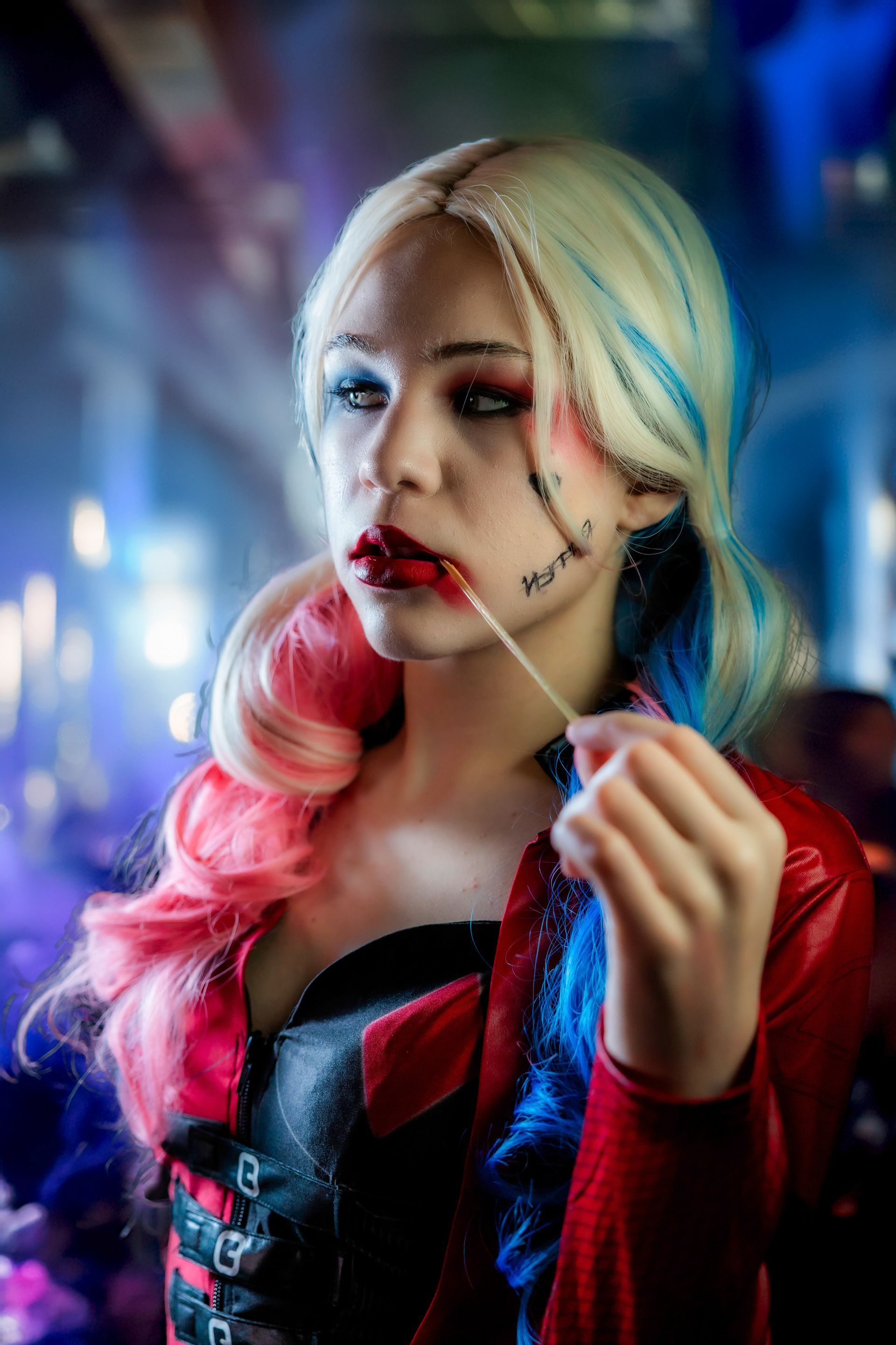 Harley Quinn cosplay with red and blue pigtails, red lipstick, holding a toothpick, and wearing a red leather jacket.