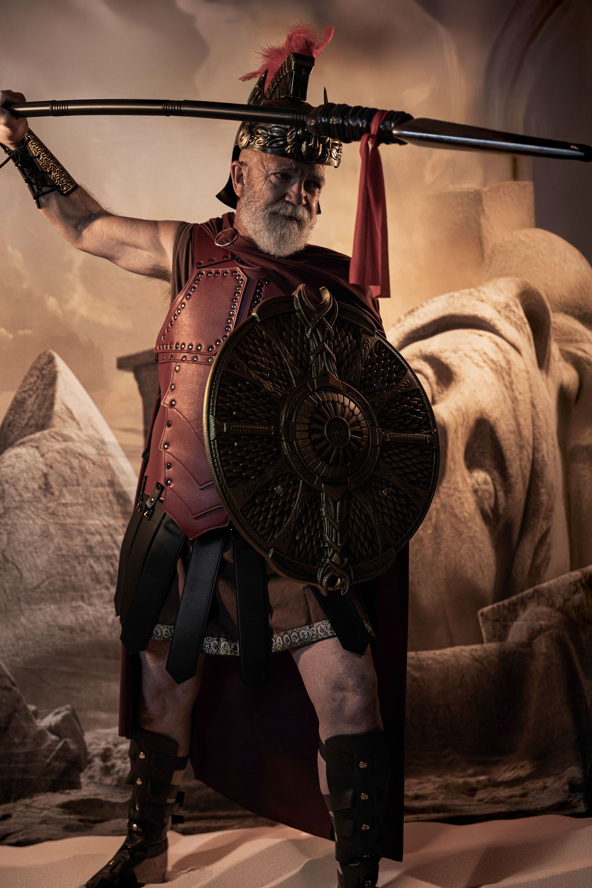 Man in Roman armor, holding spear and shield, striking a defensive pose in a desert setting.