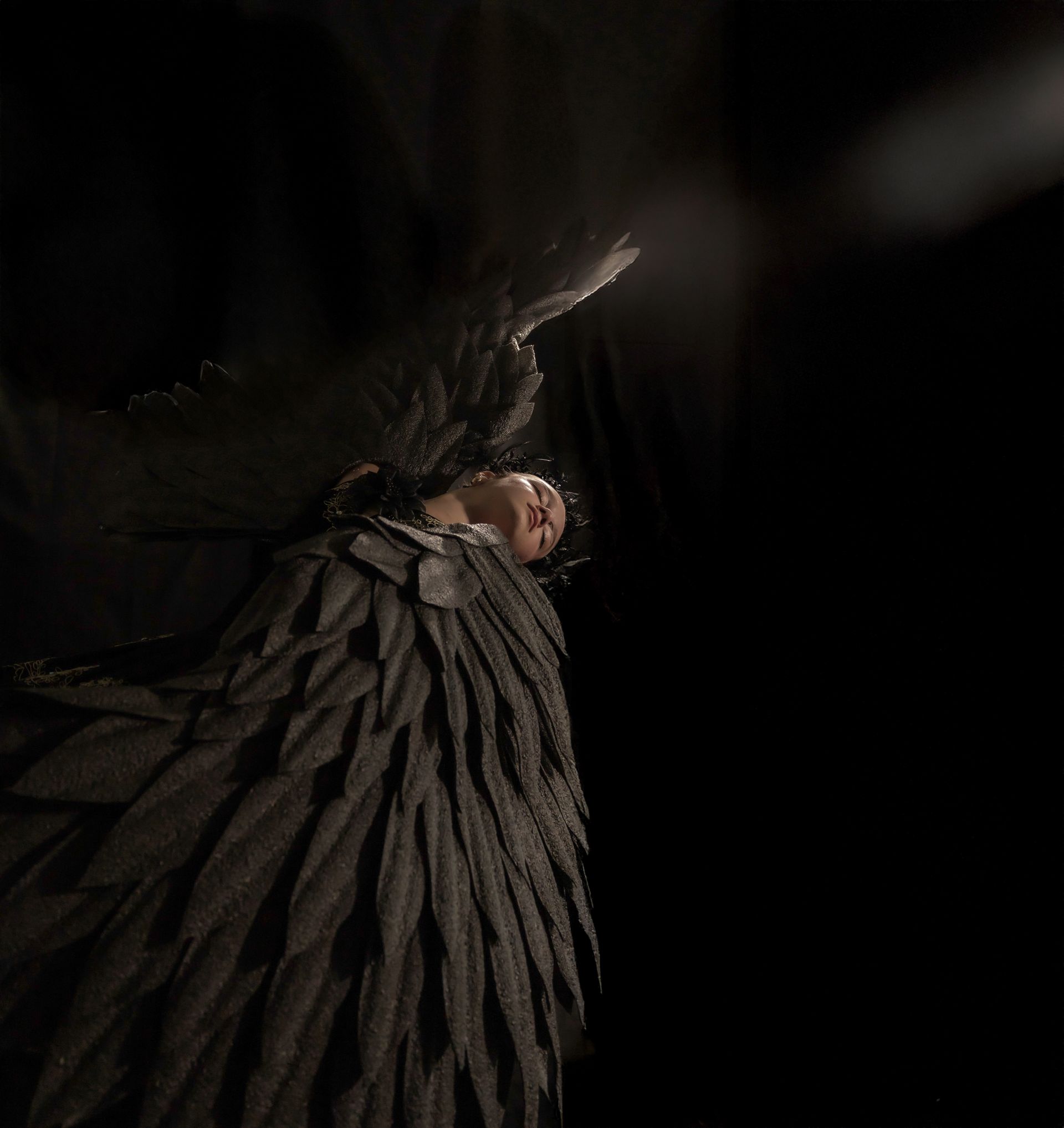 Person with black feathered wings in dark space, head tilted up.