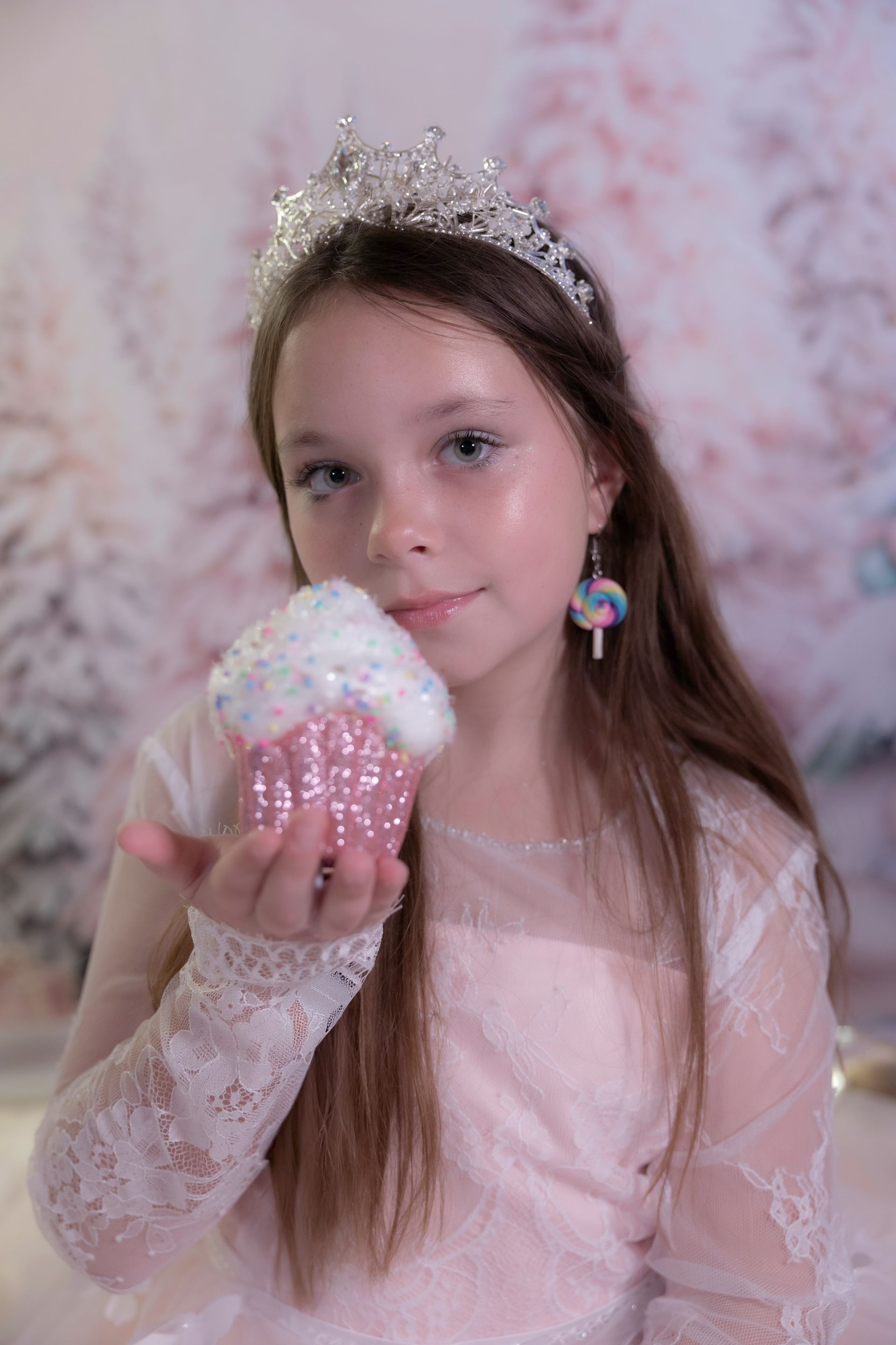 Girl in tiara and dress holding pink cupcake, festive background.