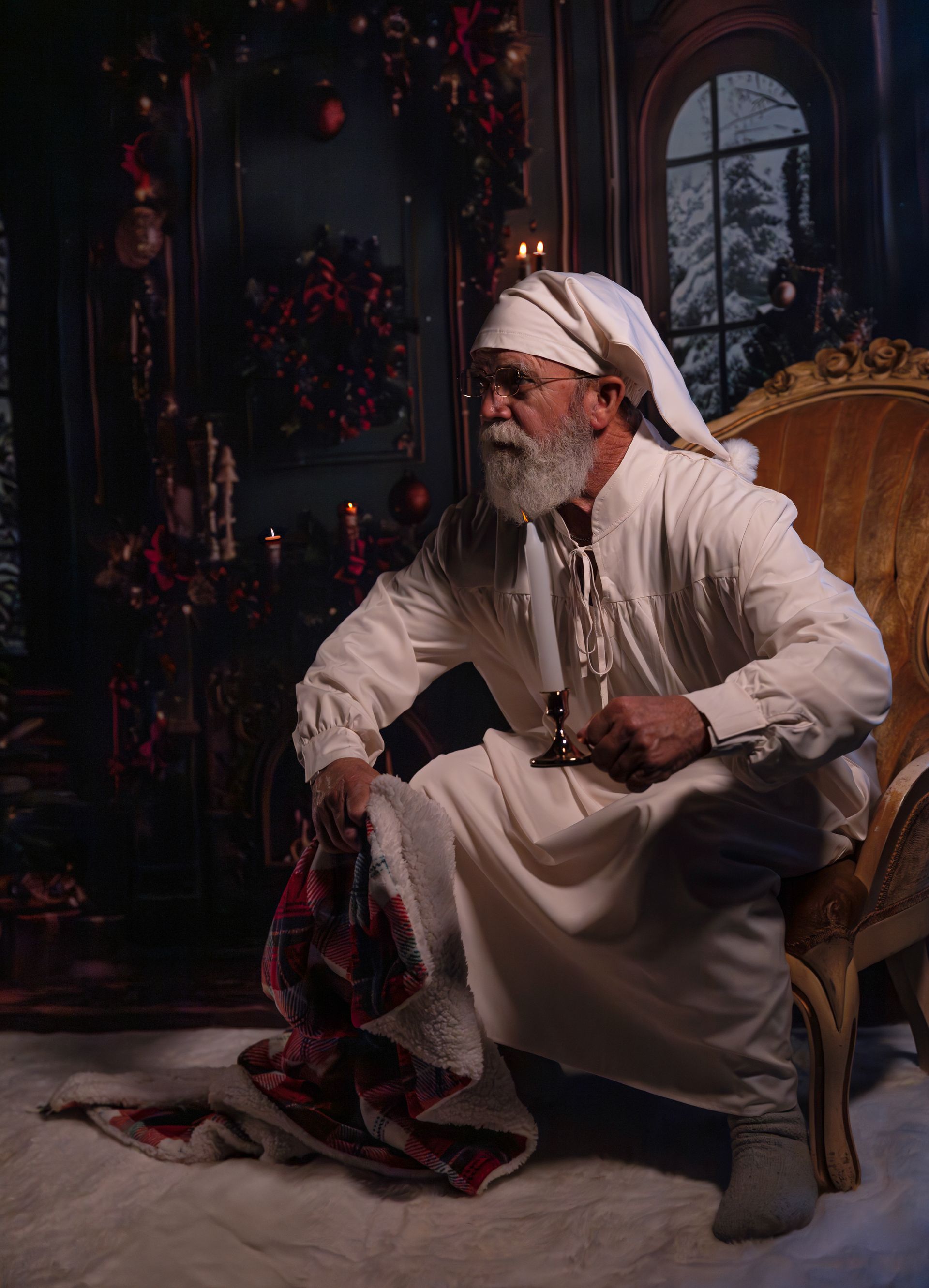 Man in nightclothes sits in ornate chair, holding a glass and plaid blanket, interior with Christmas decorations.