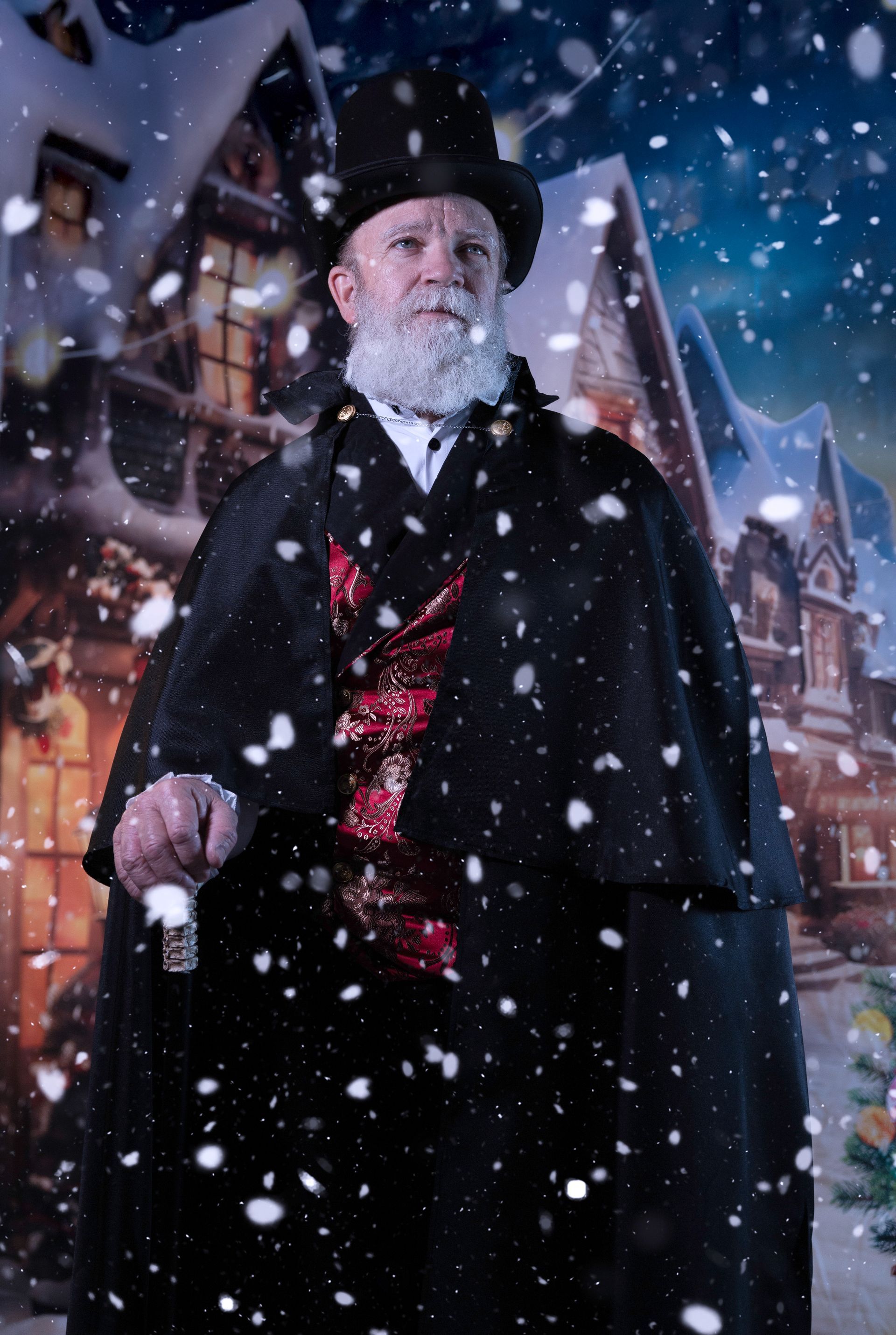 Man in Victorian attire, holding a cane, in a snowy village scene.