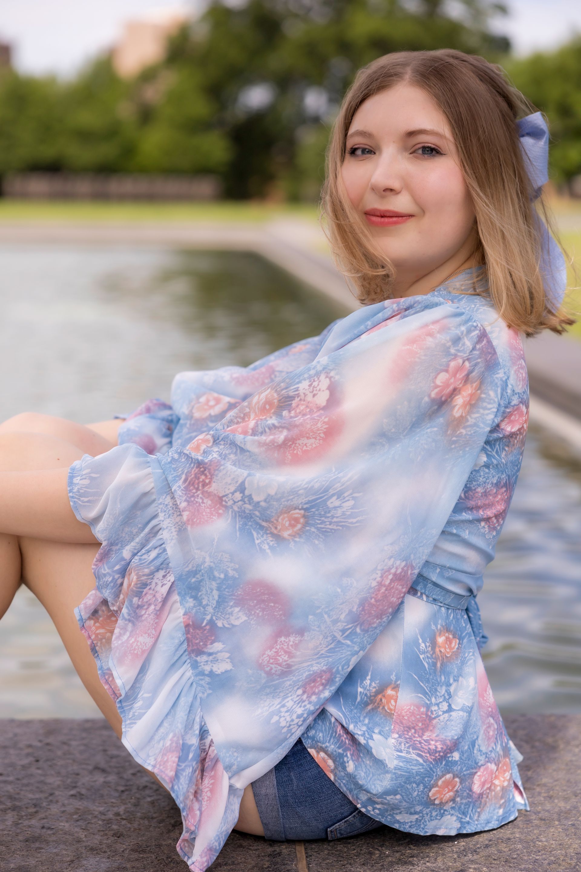 A woman in a blue floral top and shorts is sitting next to a fountain.