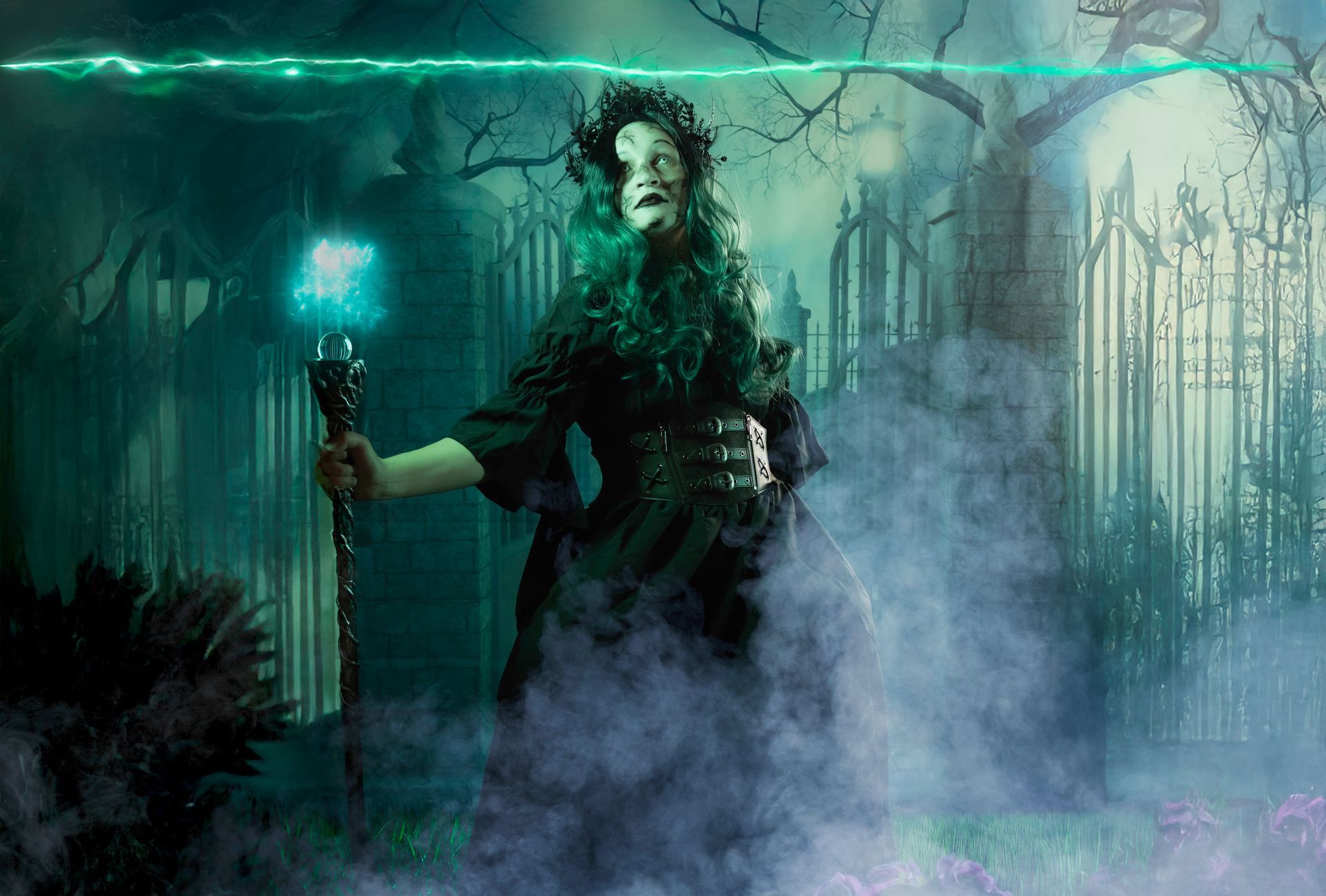 Woman in dark garb holds staff, casting green energy in graveyard.