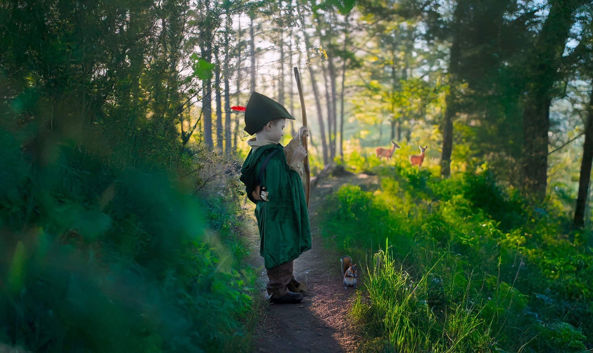 A little boy in a robin hood costume is standing on a path in the woods.