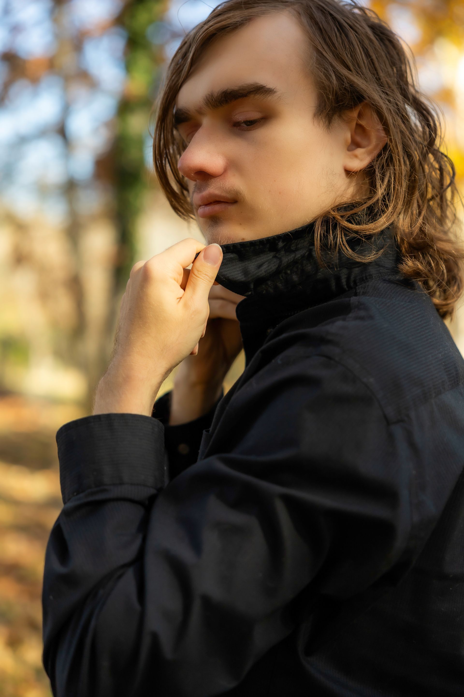 A young man with long hair is wearing a black shirt and adjusting his collar.