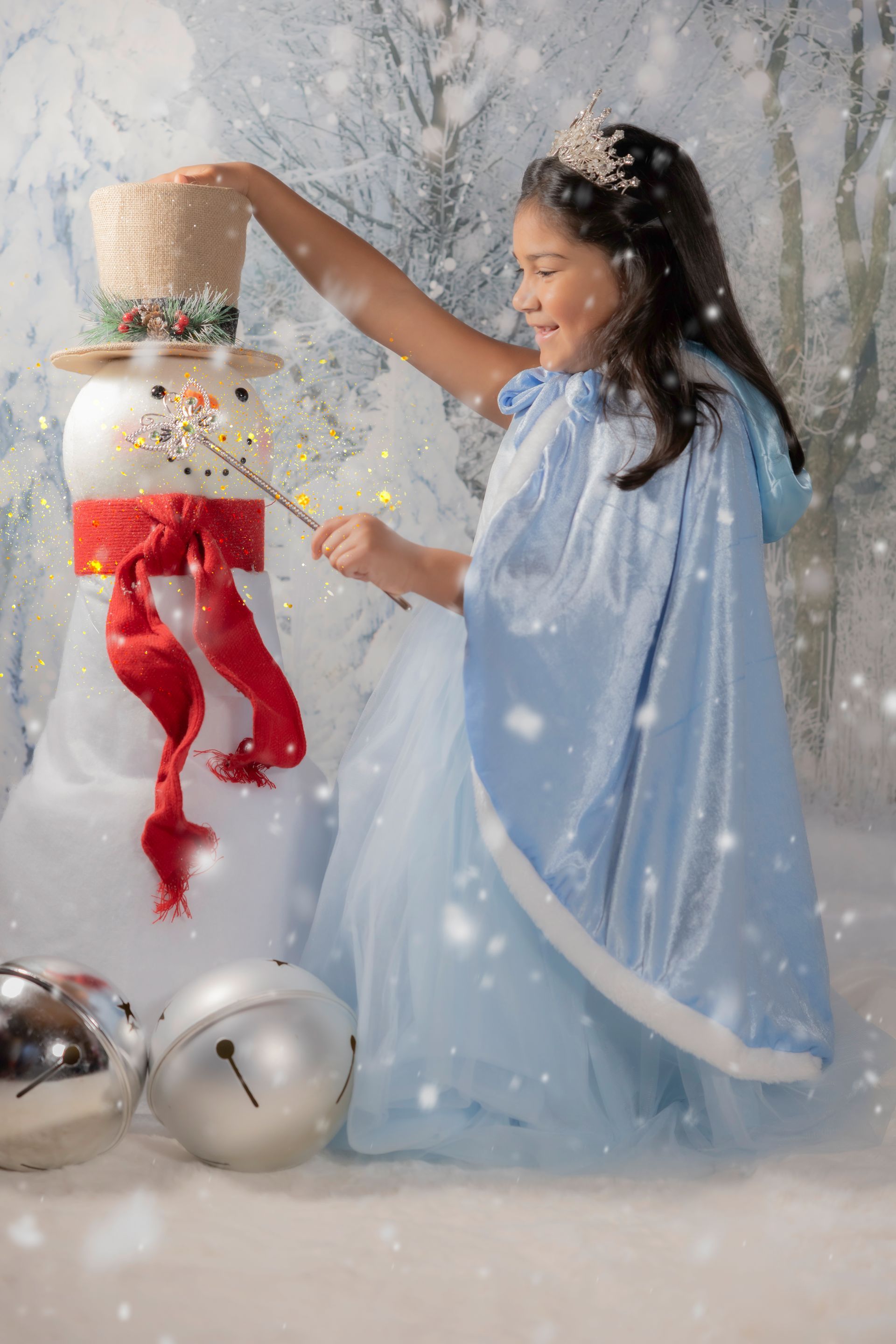 Girl in blue cape and dress, with crown, decorating a snowman with a wand; snowy background.