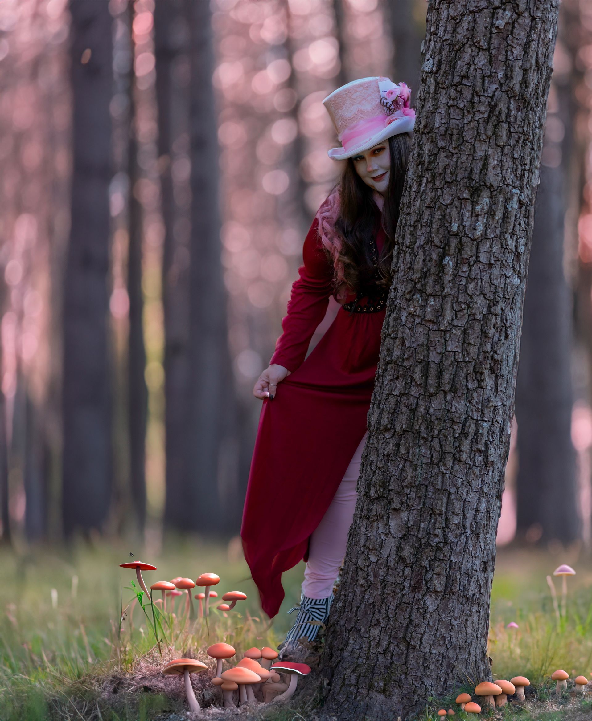 A woman in a red dress and top hat is leaning against a tree in the woods.