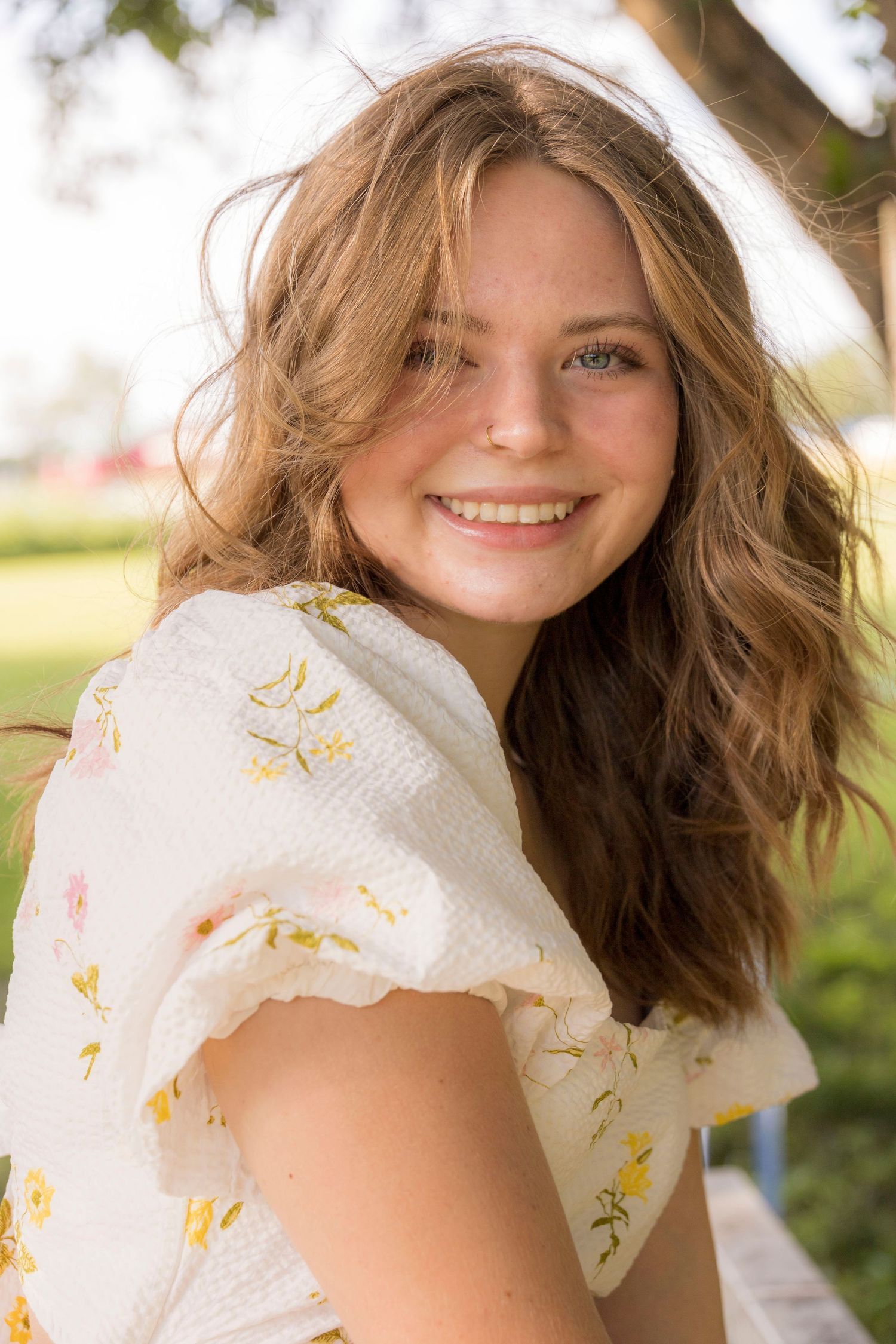 A young woman in a white dress is smiling for the camera while sitting under a tree.