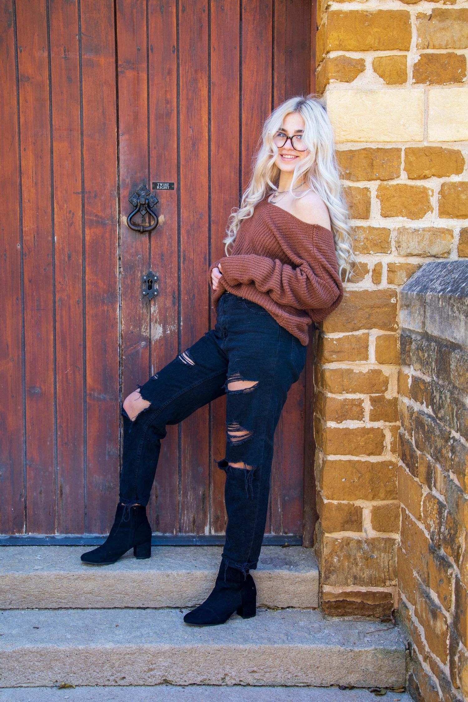 A woman in a sweater and ripped jeans is leaning against a wooden door.