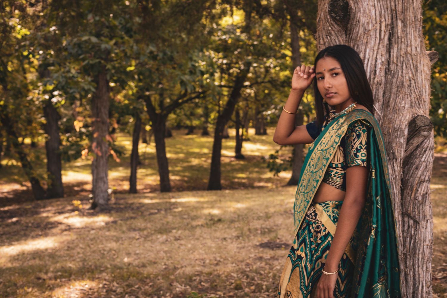 A woman in a sari is leaning against a tree in the woods.
