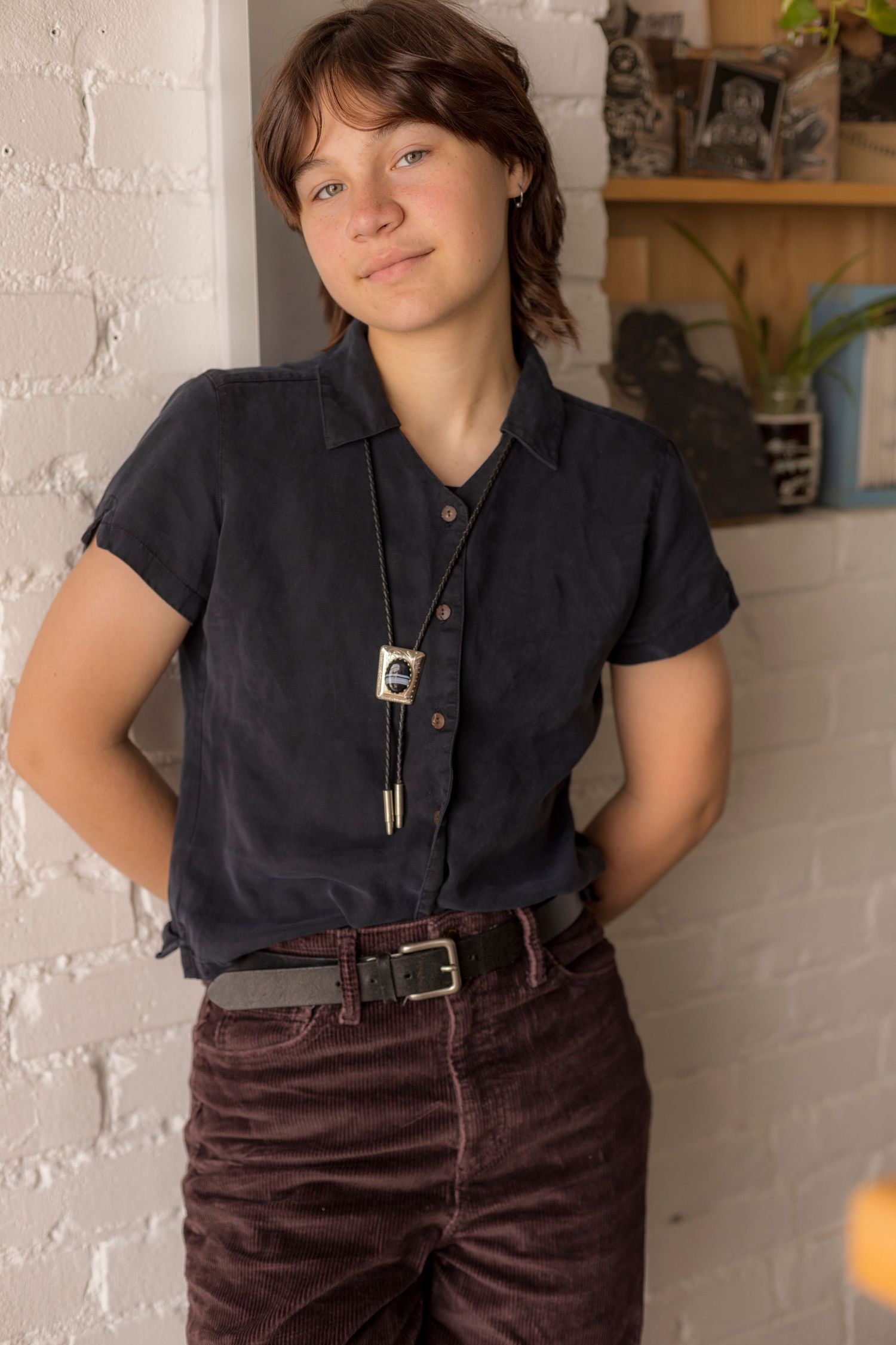 A woman wearing a black shirt and brown pants is standing in front of a brick wall.