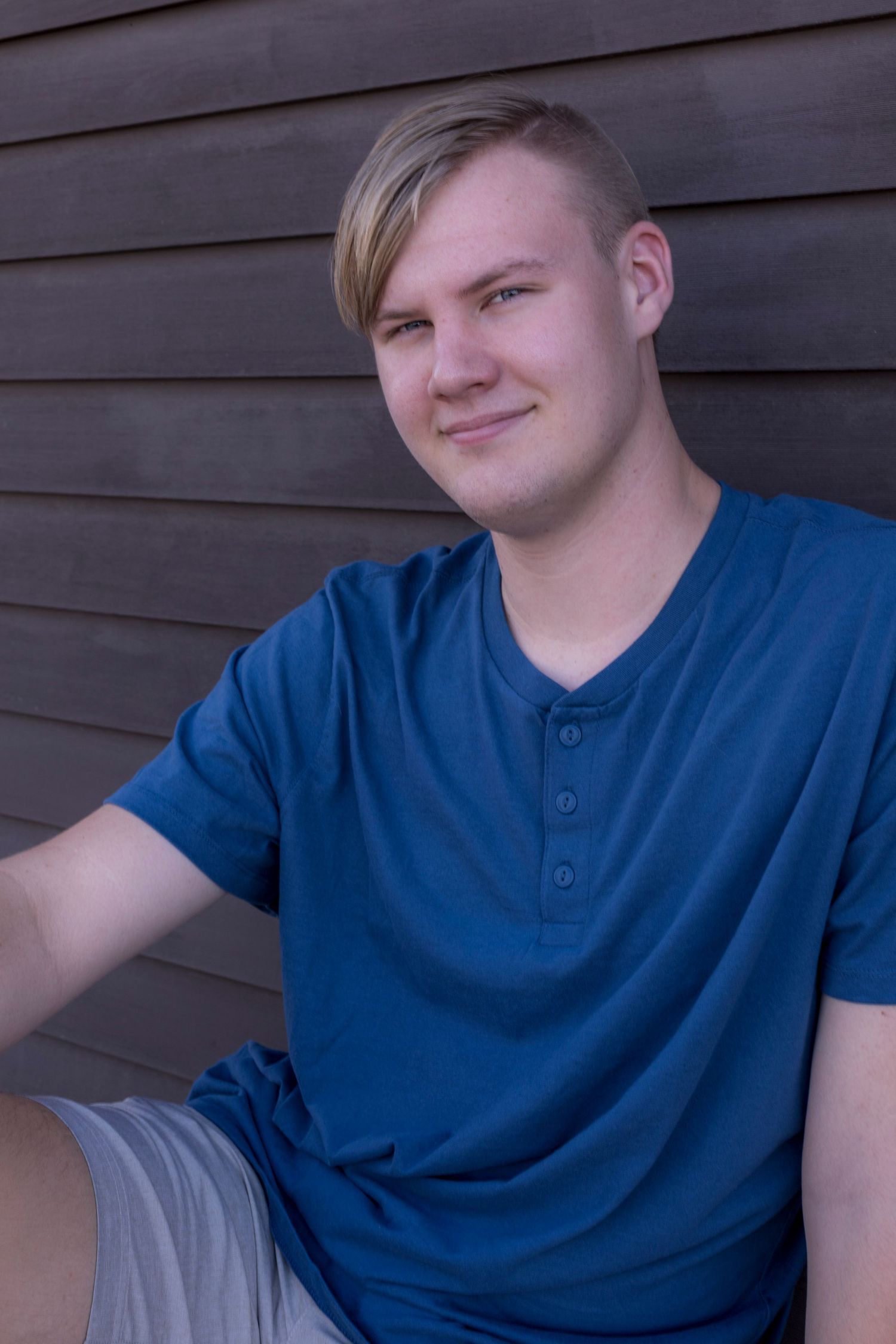 A young man in a blue shirt and shorts is sitting in front of a wooden wall.
