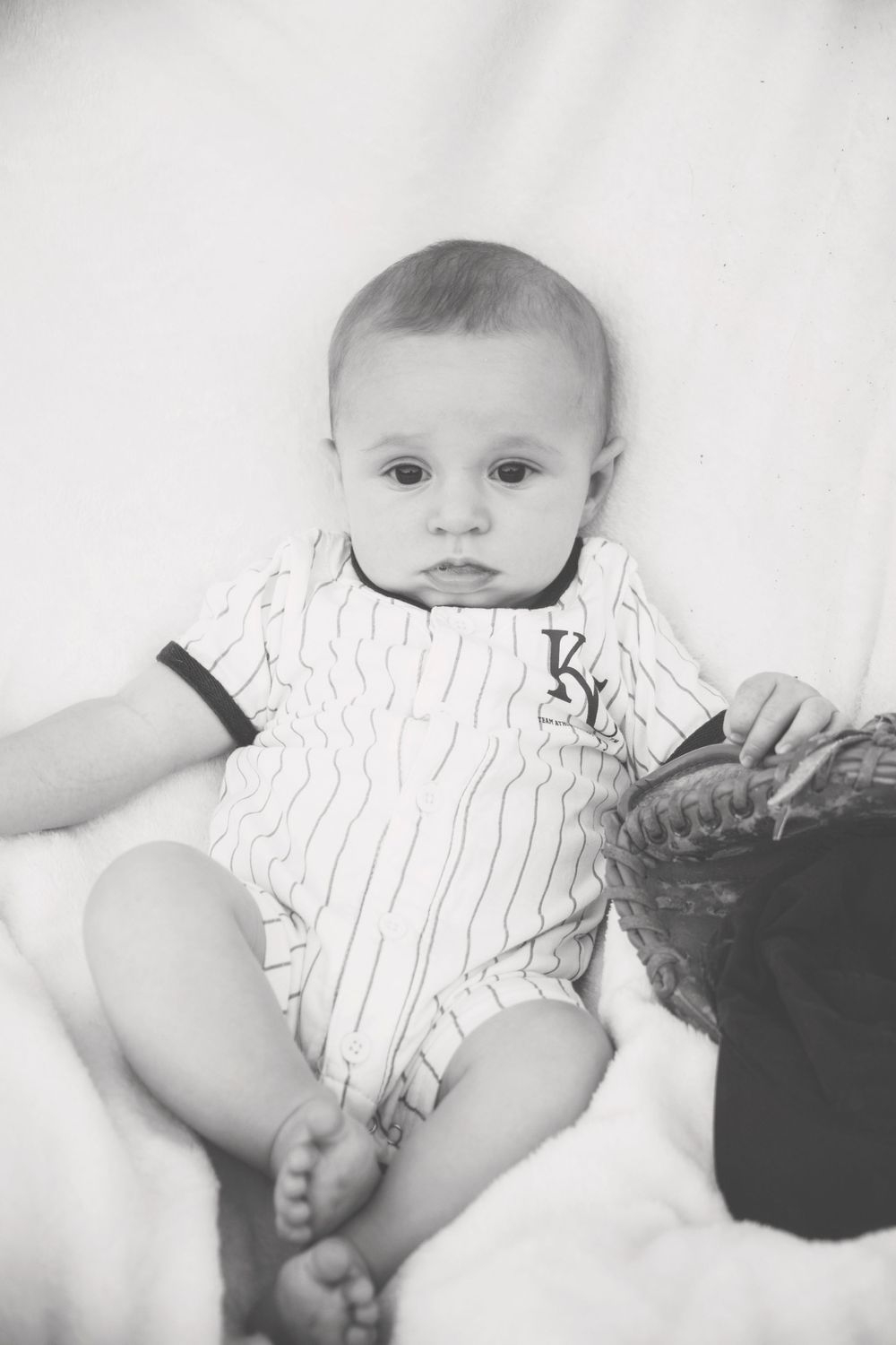 A baby in a baseball uniform is sitting on a bed.