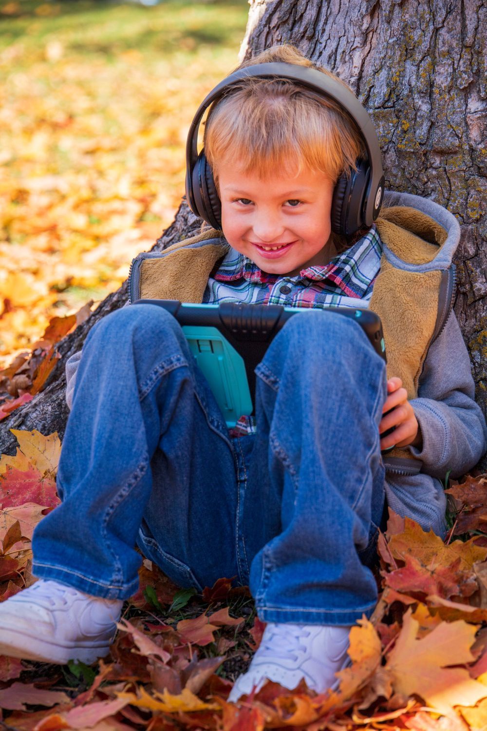 A young boy wearing headphones is sitting under a tree holding a tablet.