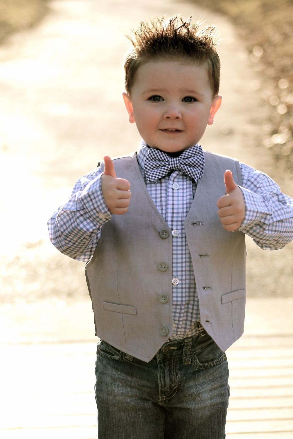 A young boy wearing a vest and bow tie is giving a thumbs up.