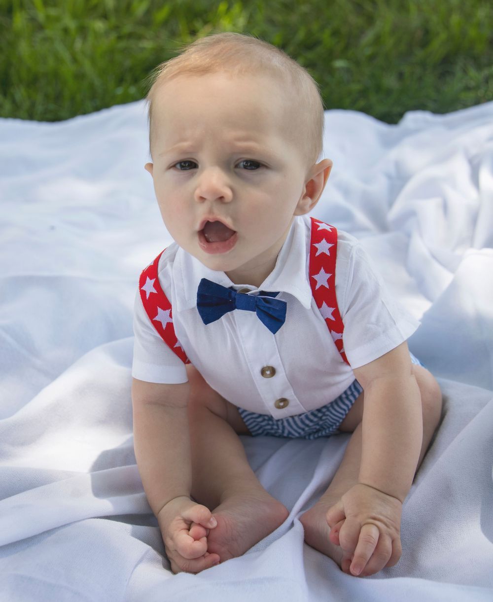 A baby wearing suspenders and a bow tie is sitting on a blanket