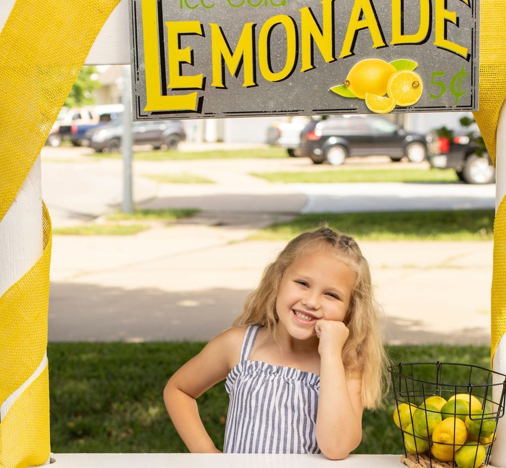 A little girl is standing in front of a lemonade stand