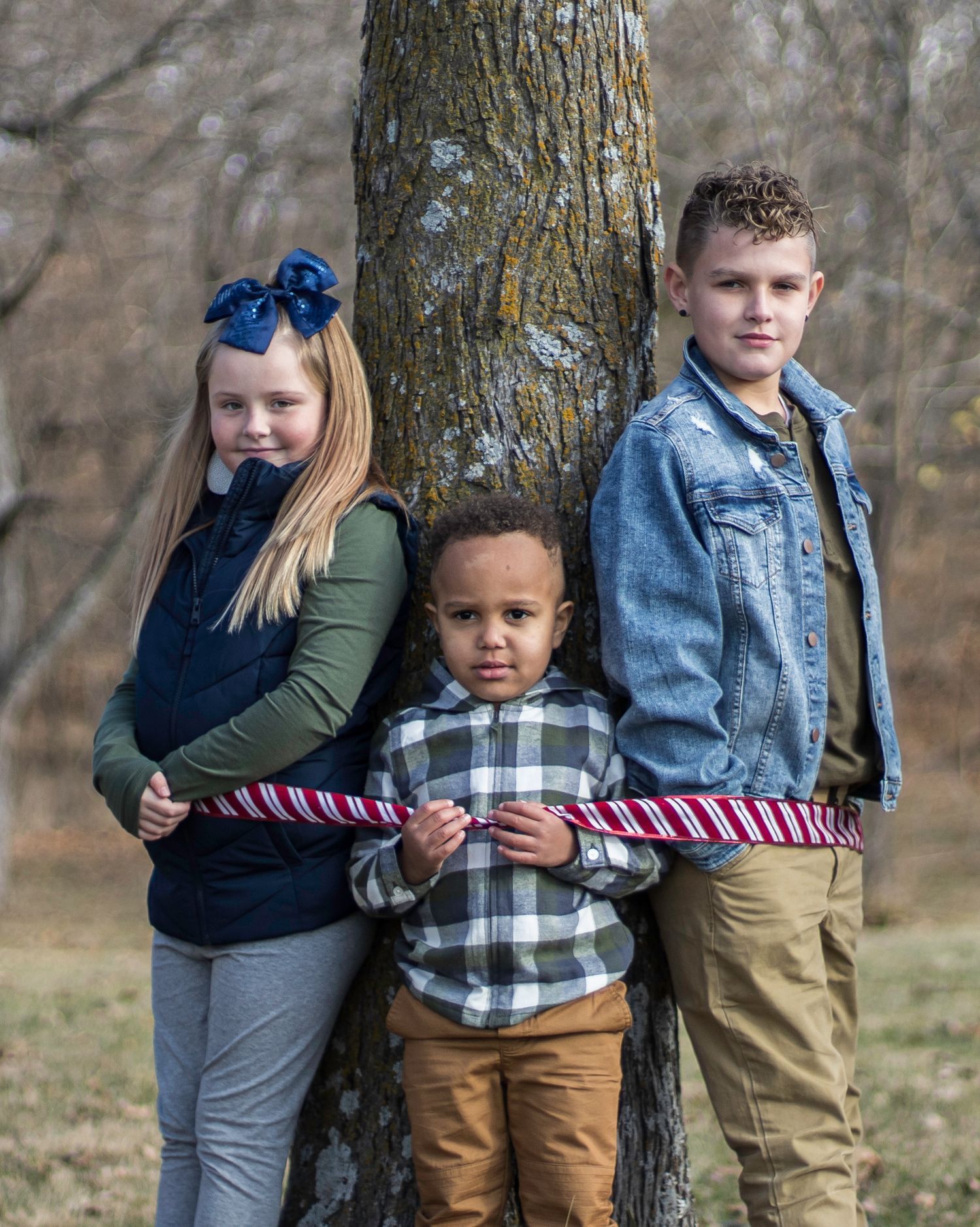 Three children are standing next to each other next to a tree.
