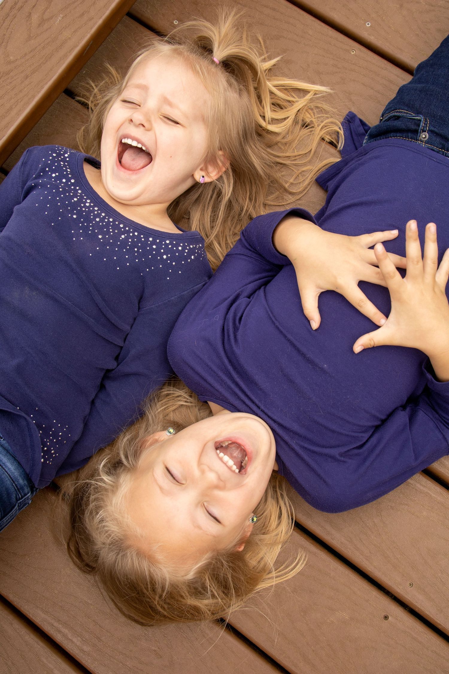 Two little girls are laying on their backs on a wooden deck and laughing.