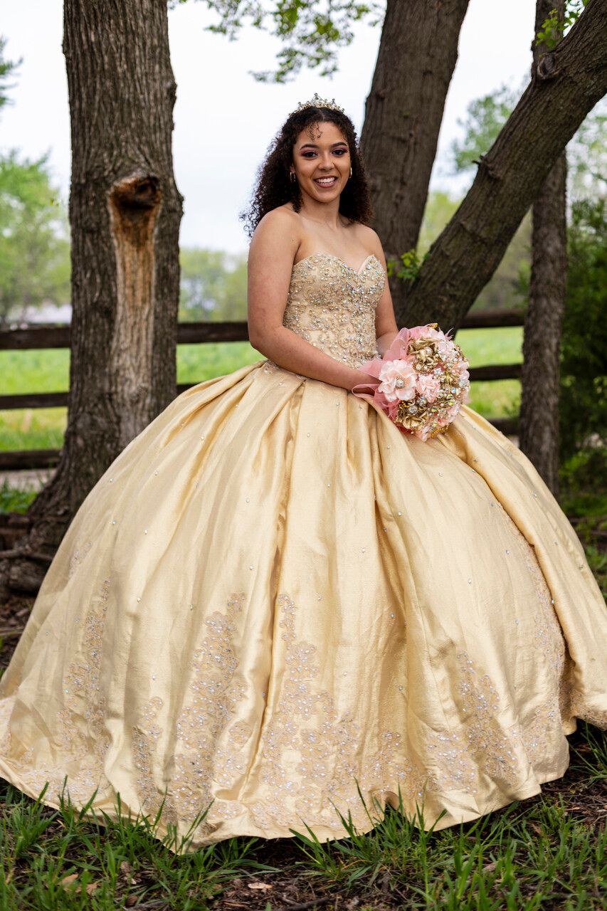 A woman in a gold ball gown is standing in front of a tree holding a bouquet of flowers.
