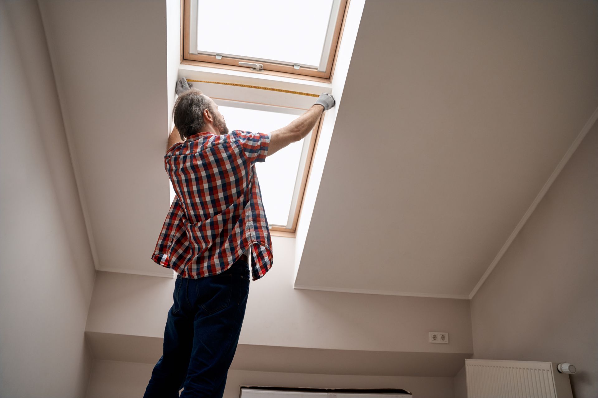 A man holding a measuring tape stands beside a skylight window.