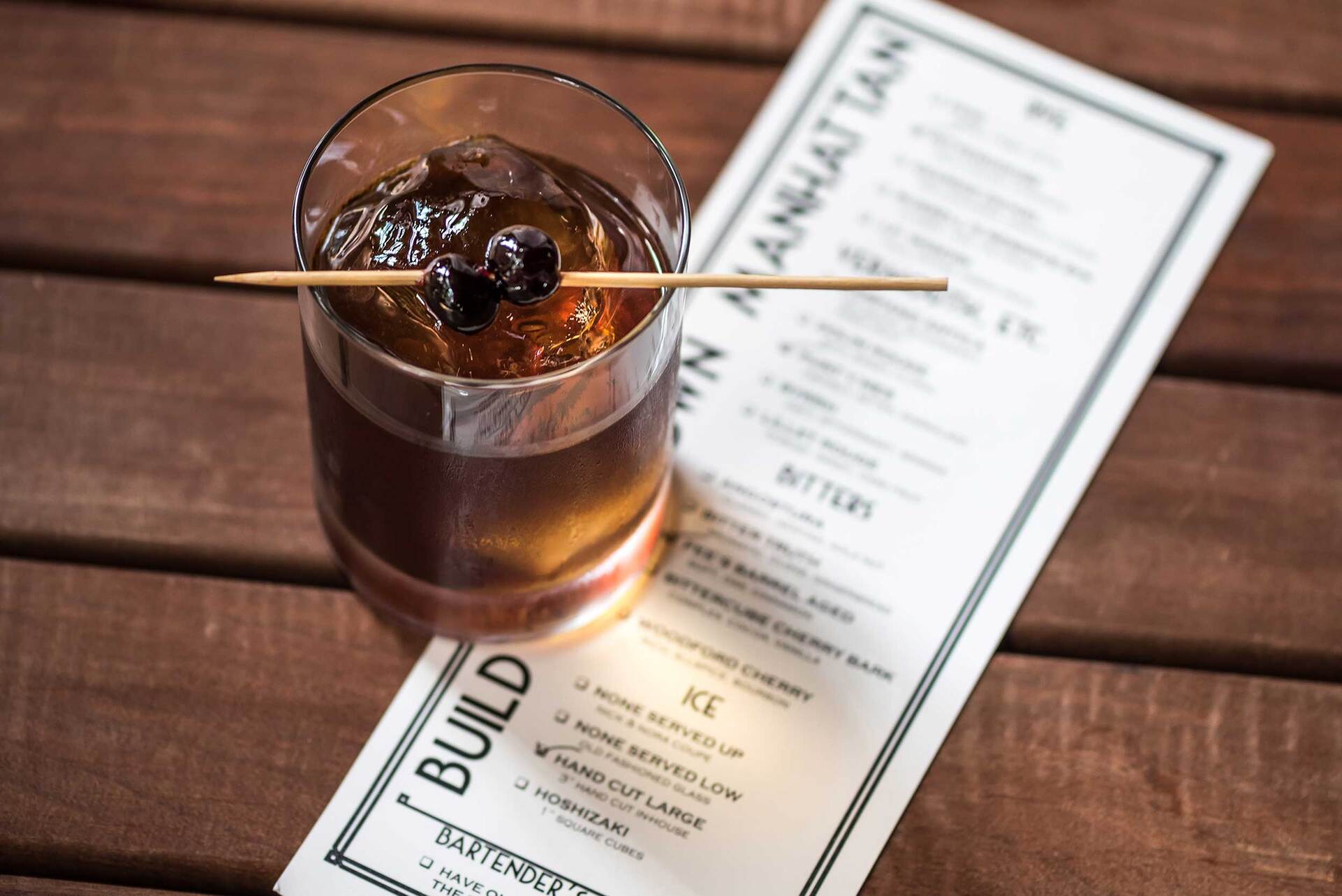 A drink with a cherry on a toothpick sits on a wooden table next to a menu