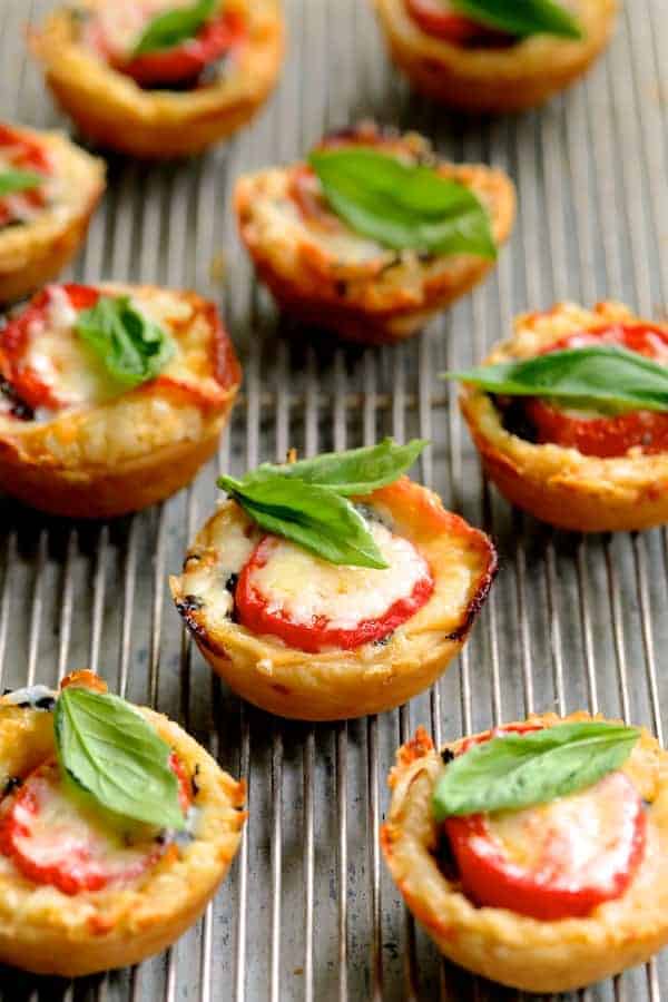 A bunch of mini pizzas with tomatoes and basil on a metal tray.