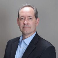 Dan Sharp wearing a blue shirt and black blazer posing for a headshot against a gray background.