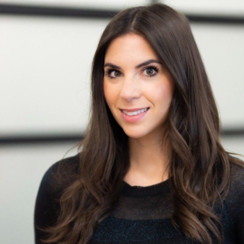 Woman with long brown hair smiles at the camera, wearing a black top in an office setting.