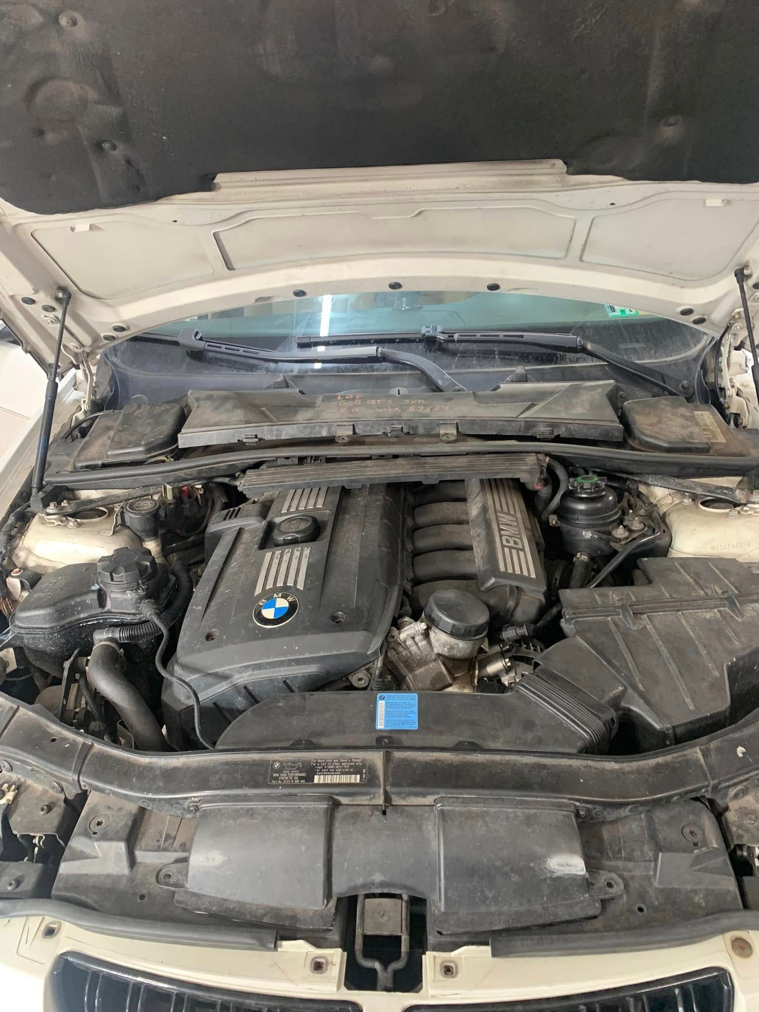 Open car hood revealing a BMW engine bay. The engine is black with a blue emblem, surrounded by other car components.