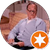 Older man in glasses and light blue shirt, seated on a couch, looking down, with a star badge.