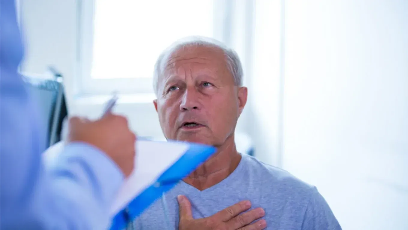Una persona mayor sentada en una consulta médica, con la mano sobre el pecho mientras habla con un profesional sanitario.