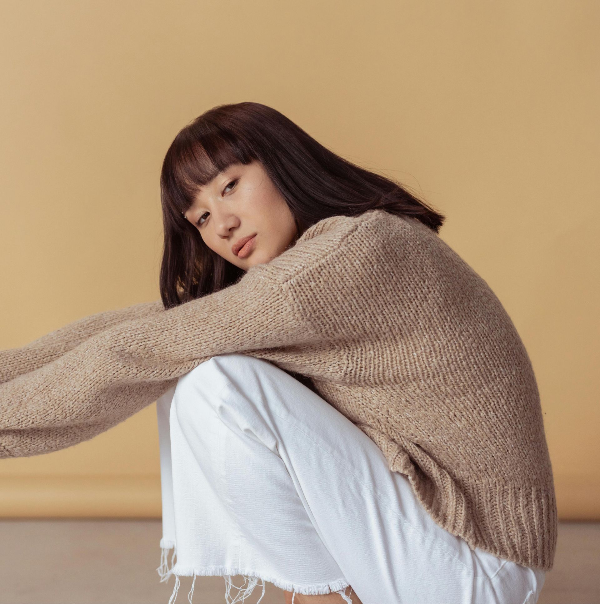 A woman wearing a tan sweater and white pants is squatting down