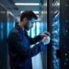 Man working on server equipment in a data center, focused, wearing a dark blue jacket.