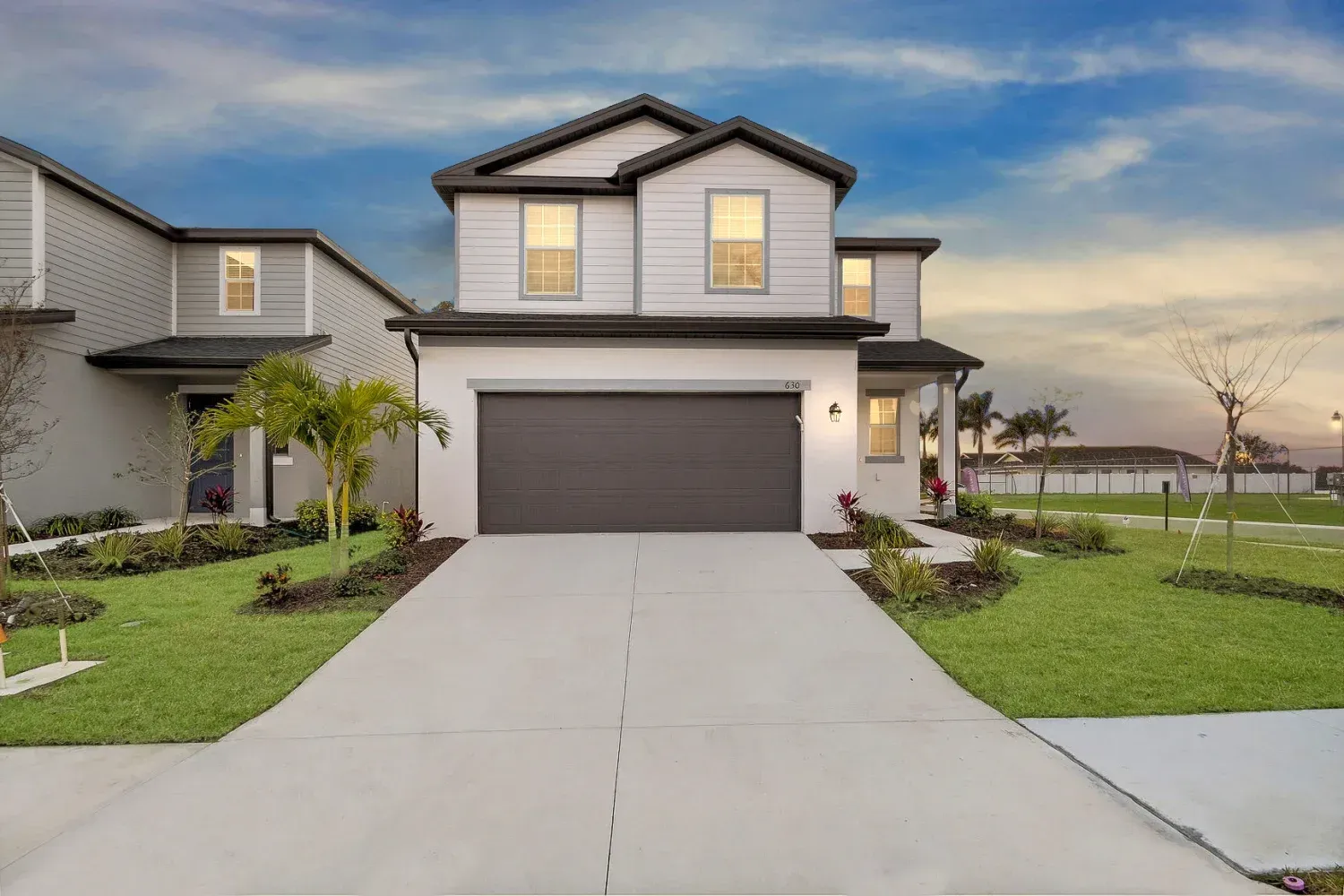 A two-story suburban house with white siding, a dark roof, and a large concrete driveway under a sunset sky.