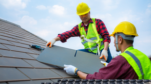 Two workers in safety vests and helmets install a dark panel on a tiled roof.