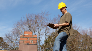 Construction worker in yellow hard hat on roof, inspecting chimney with clipboard. Blue sky background.