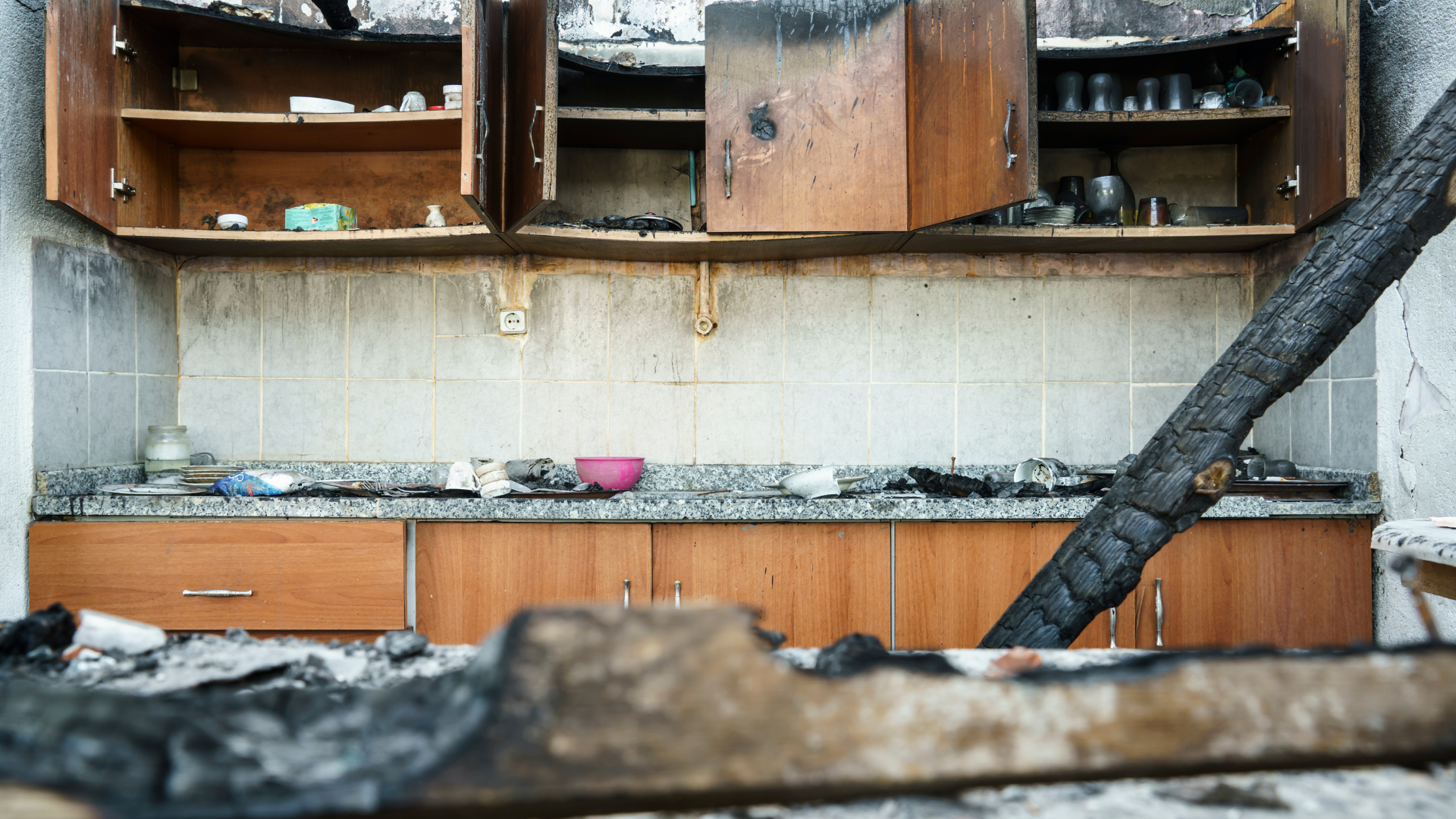 Burned kitchen with charred cabinets, debris, and structural damage from a fire.