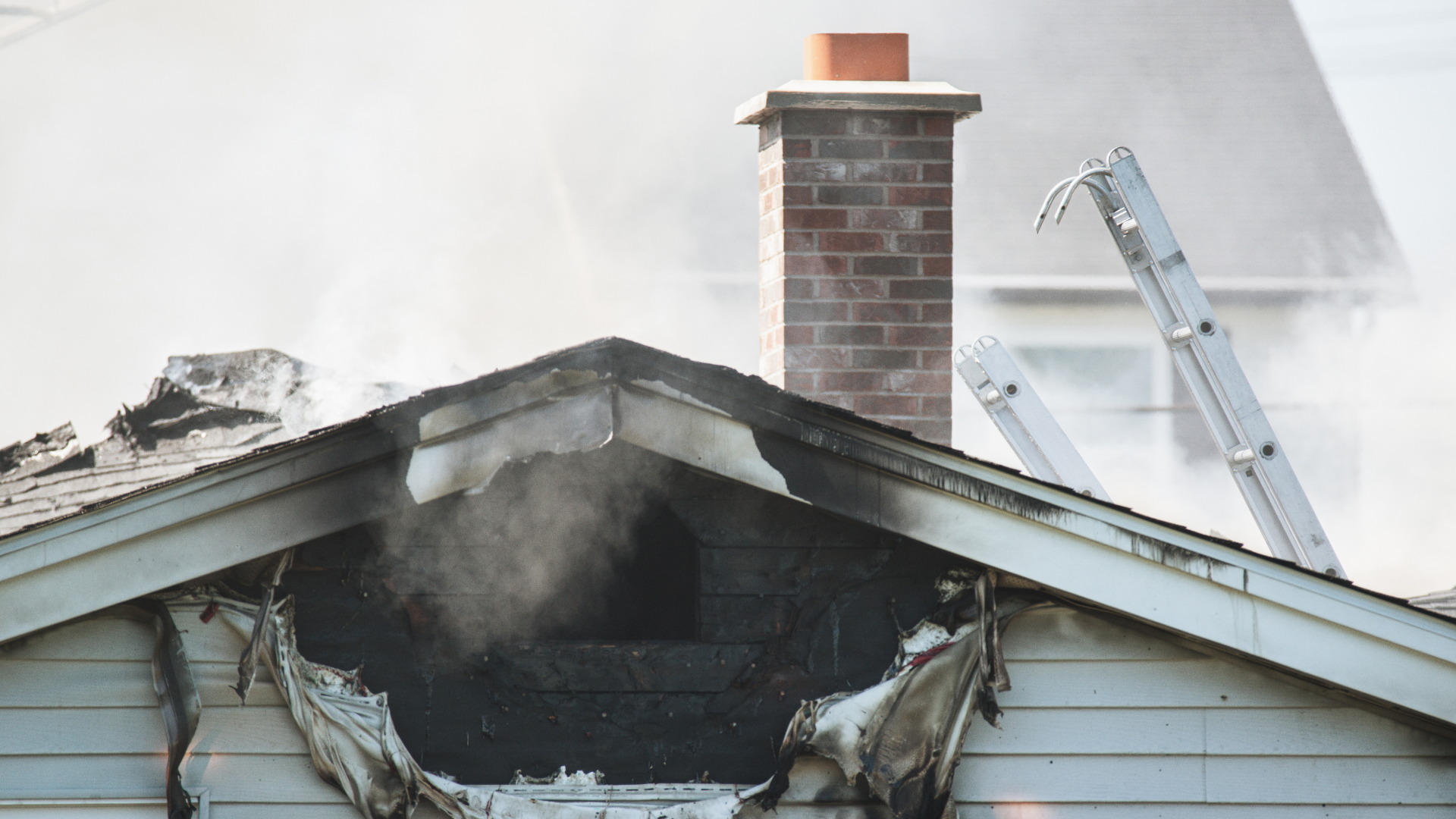A house roof with a large hole, smoke billowing. Brick chimney and ladder visible.