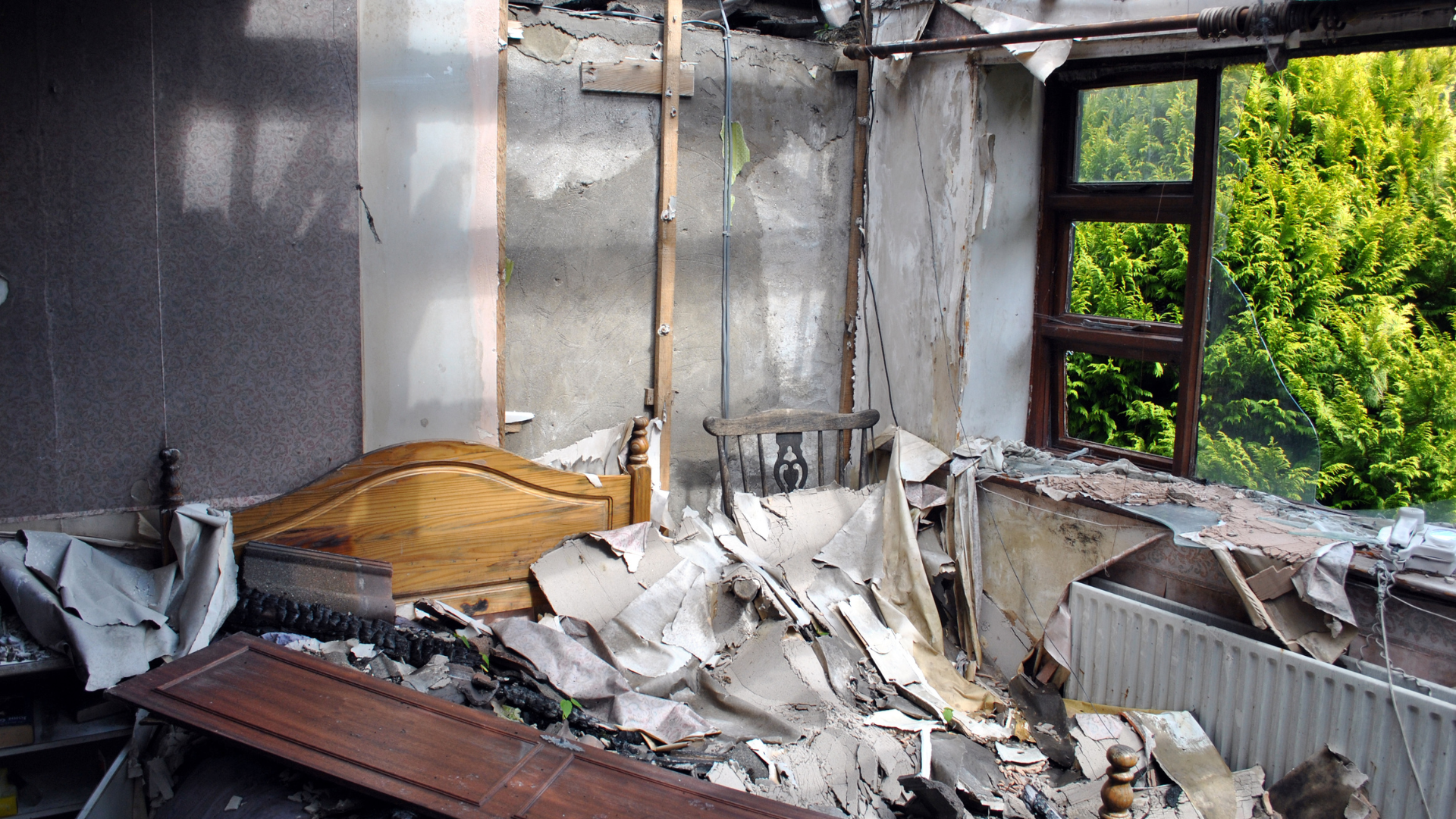 Room damaged by fire, debris scattered, bed frame visible, window with green foliage.