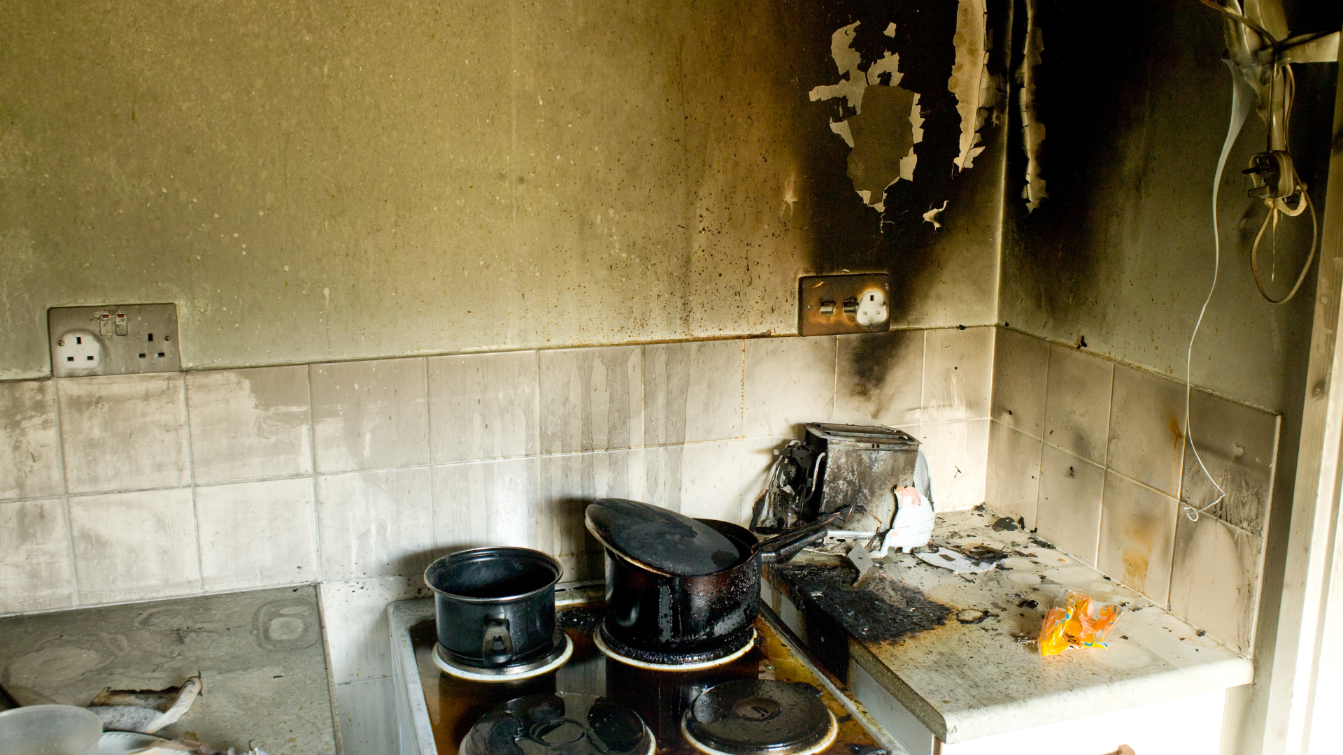 Kitchen interior damaged by fire, showing soot-covered walls, burnt appliances, and debris.