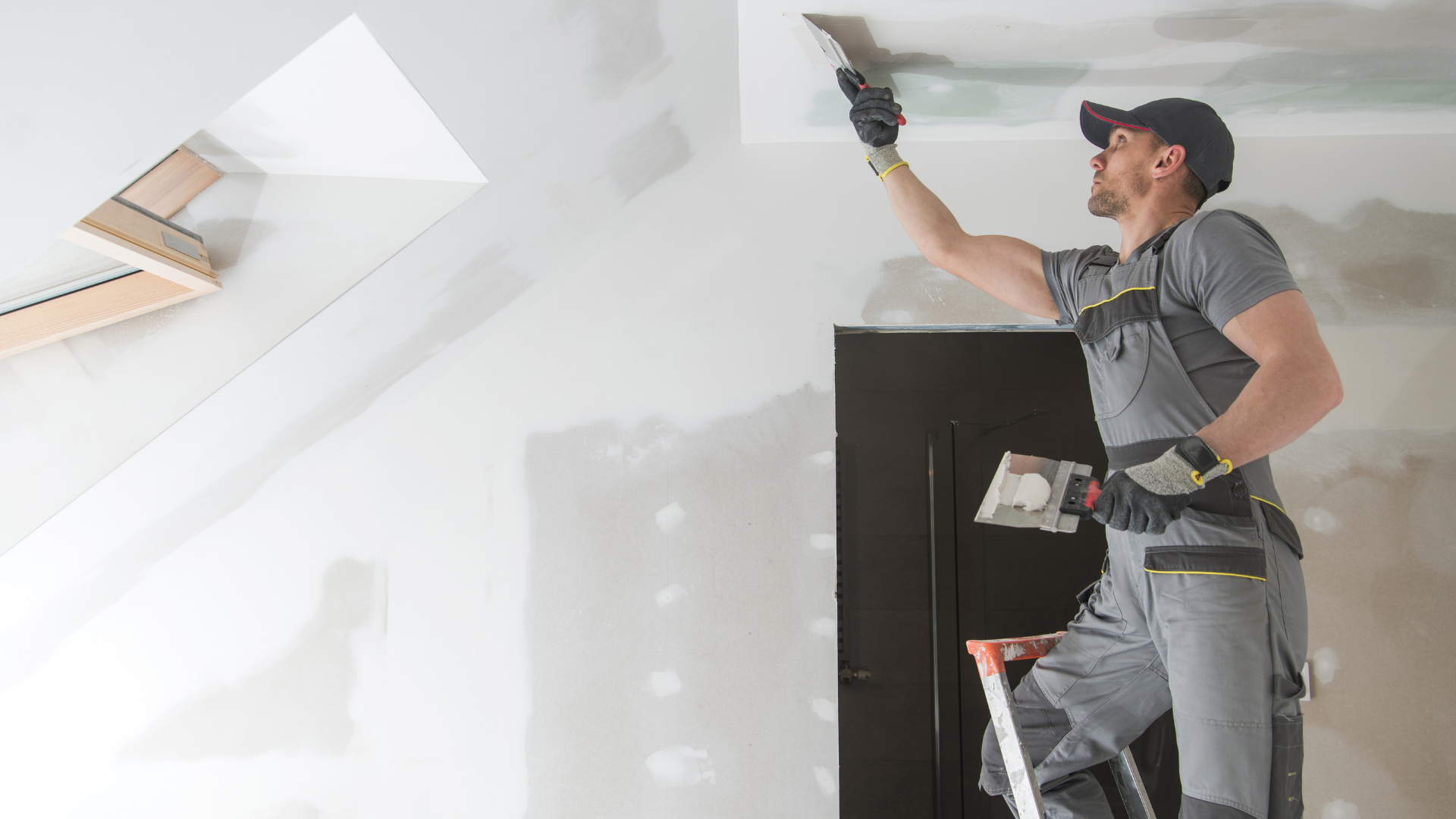 Construction worker on a ladder, using a trowel to apply drywall mud to a ceiling.