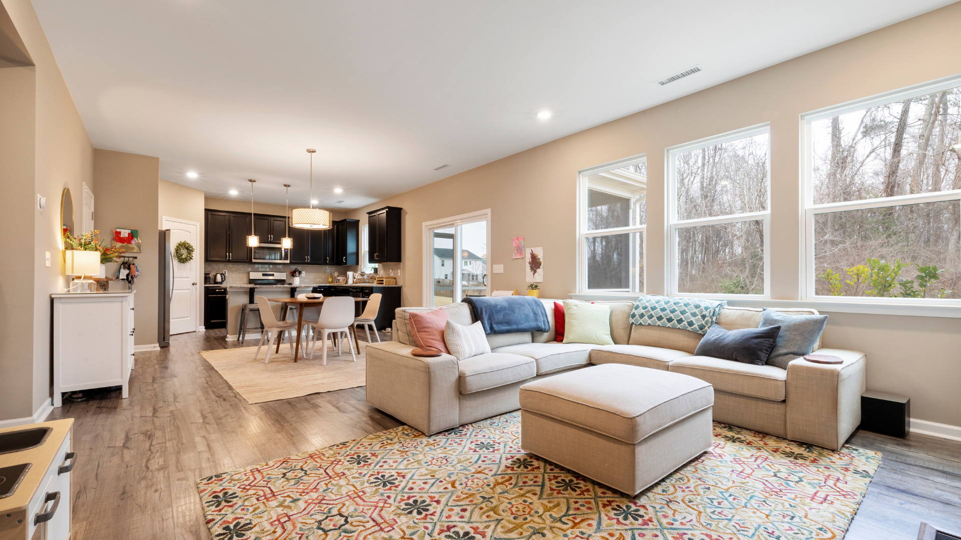 Open-concept living space with a sectional sofa, rug, and kitchen in the background, next to large windows.