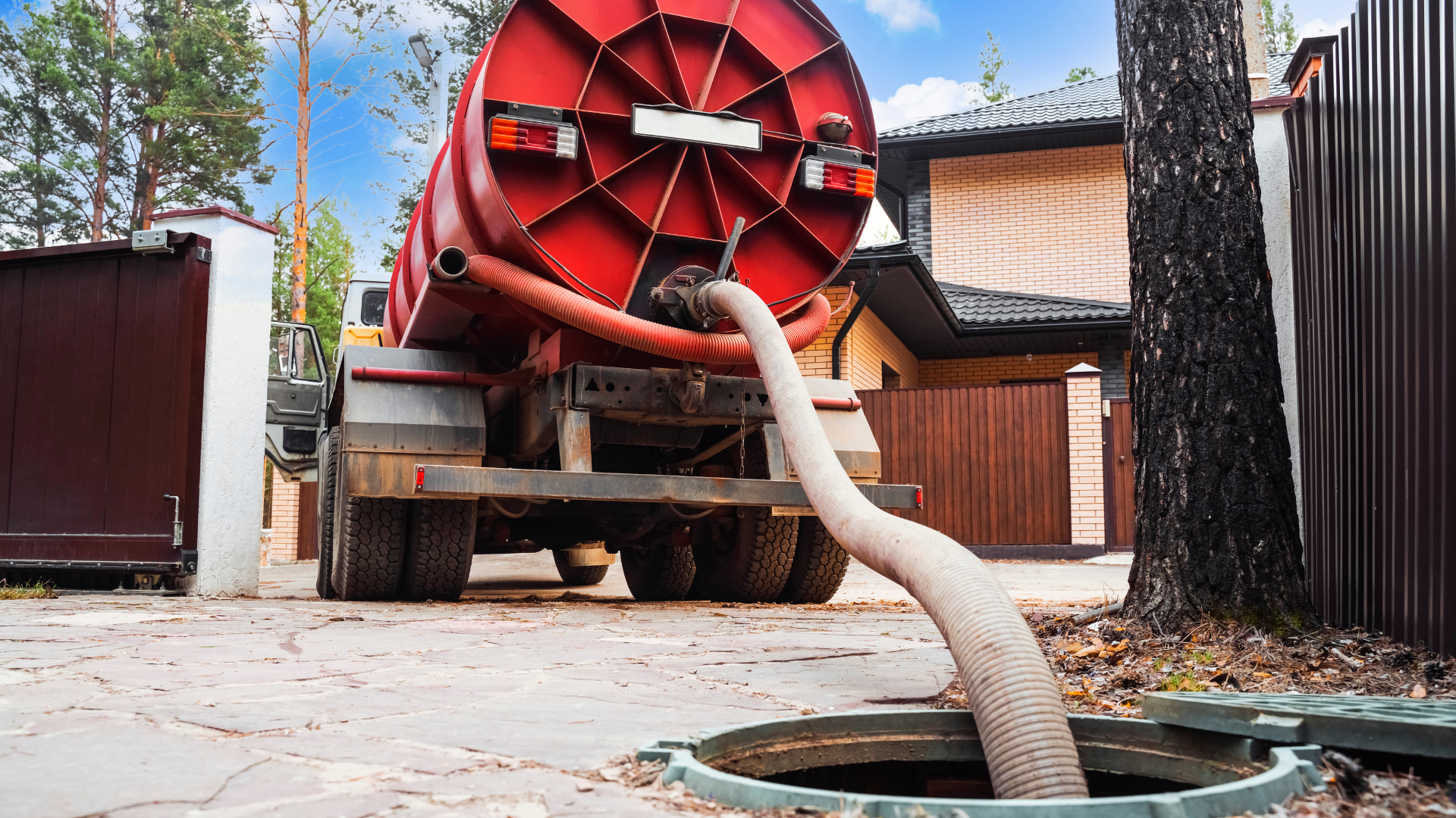 Red septic tank truck emptying a tank via a hose into a ground-level access hole at a residential property.