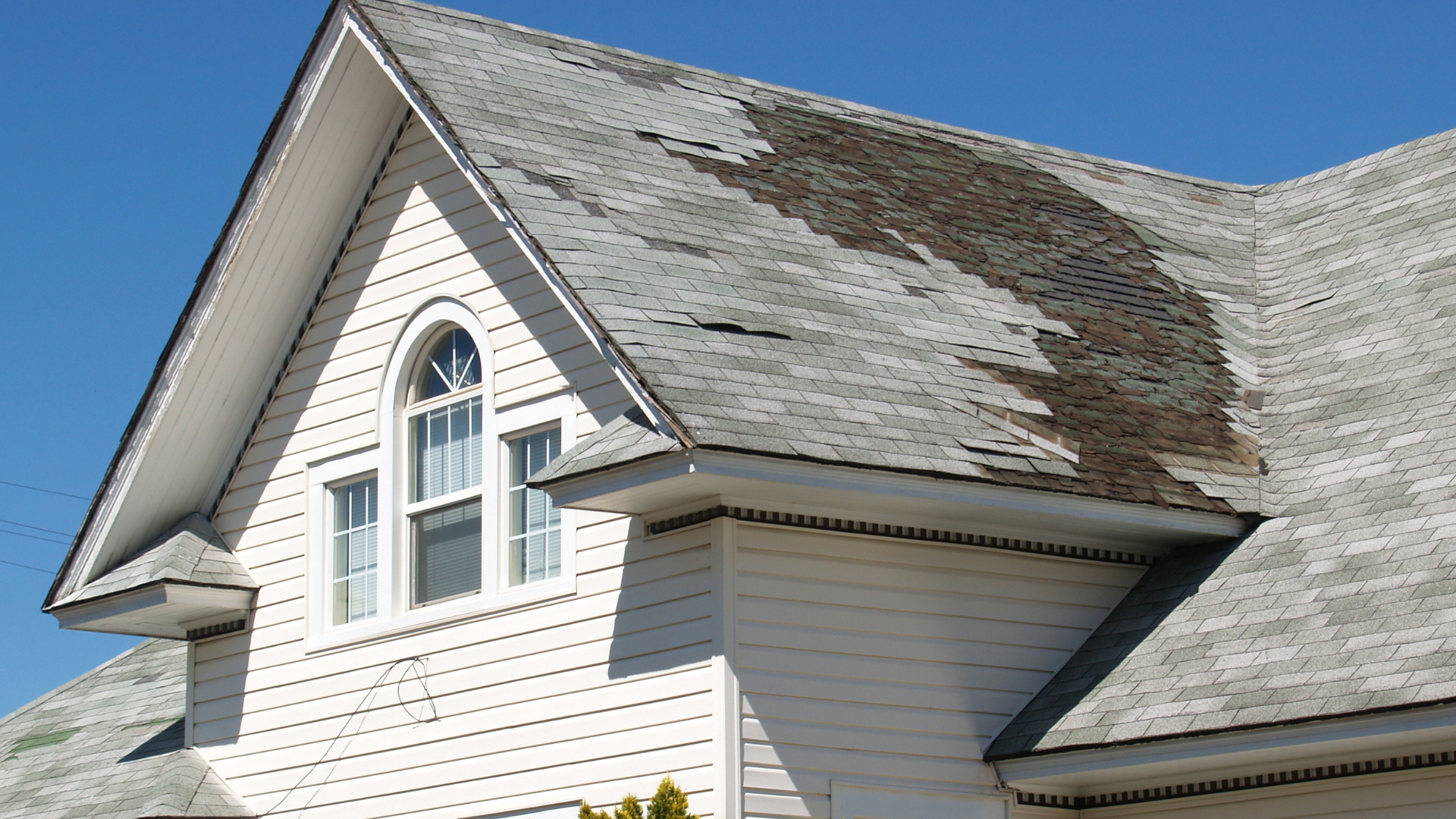 Damaged roof on a white house, shingles peeling away, against a blue sky.