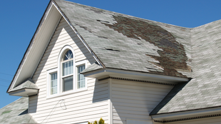 Damaged roof on a white house, shingles peeling away, against a blue sky.
