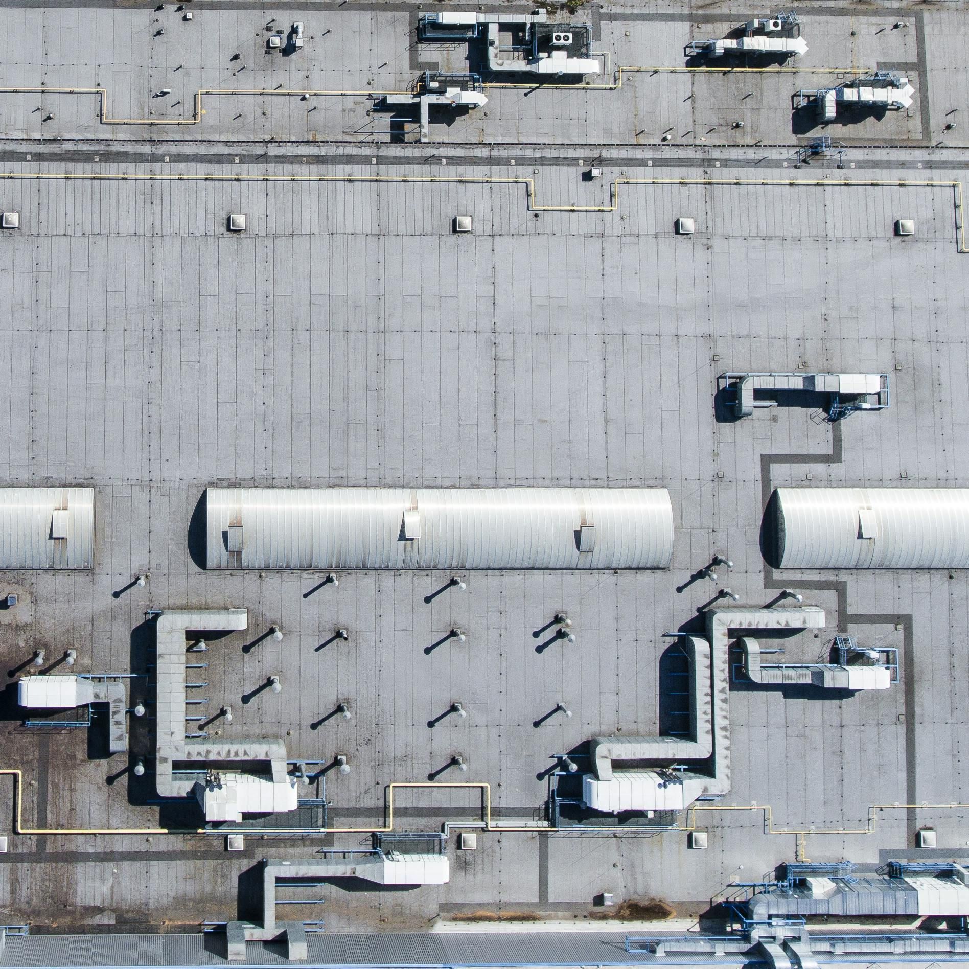 Overhead view of a gray industrial building rooftop, with ventilation, pipes, and other structures.