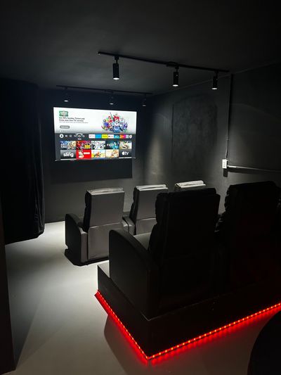 A dark home theater room with gray recliners facing a large TV. Red lights illuminate the base of the seating.